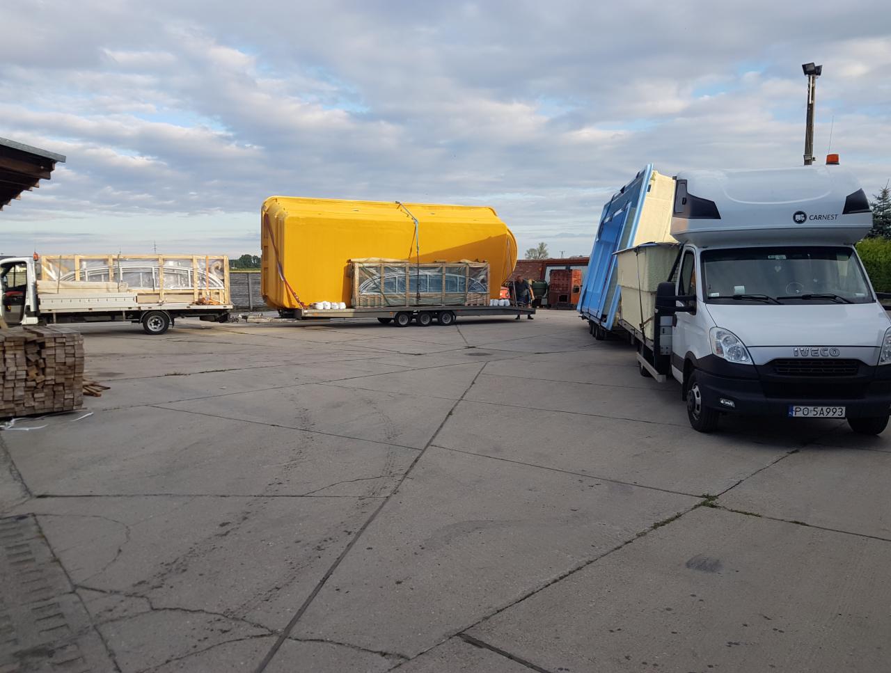 Trzy samochody dostawcze na placu, załadowane oknami i elementami konstrukcyjnymi, zabezpieczonymi folią i drewnianymi ramami, gotowe do przewozu.