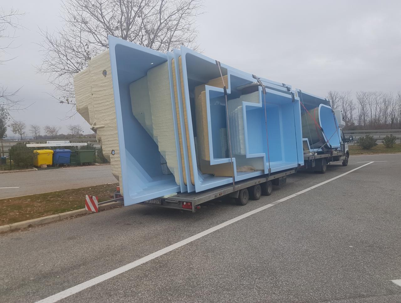 Ciężarówka przewozi kilka gotowych, niebieskich basenów z izolacją termiczną, zabezpieczonych pasami, na tle szarej drogi i drzew bez liści.