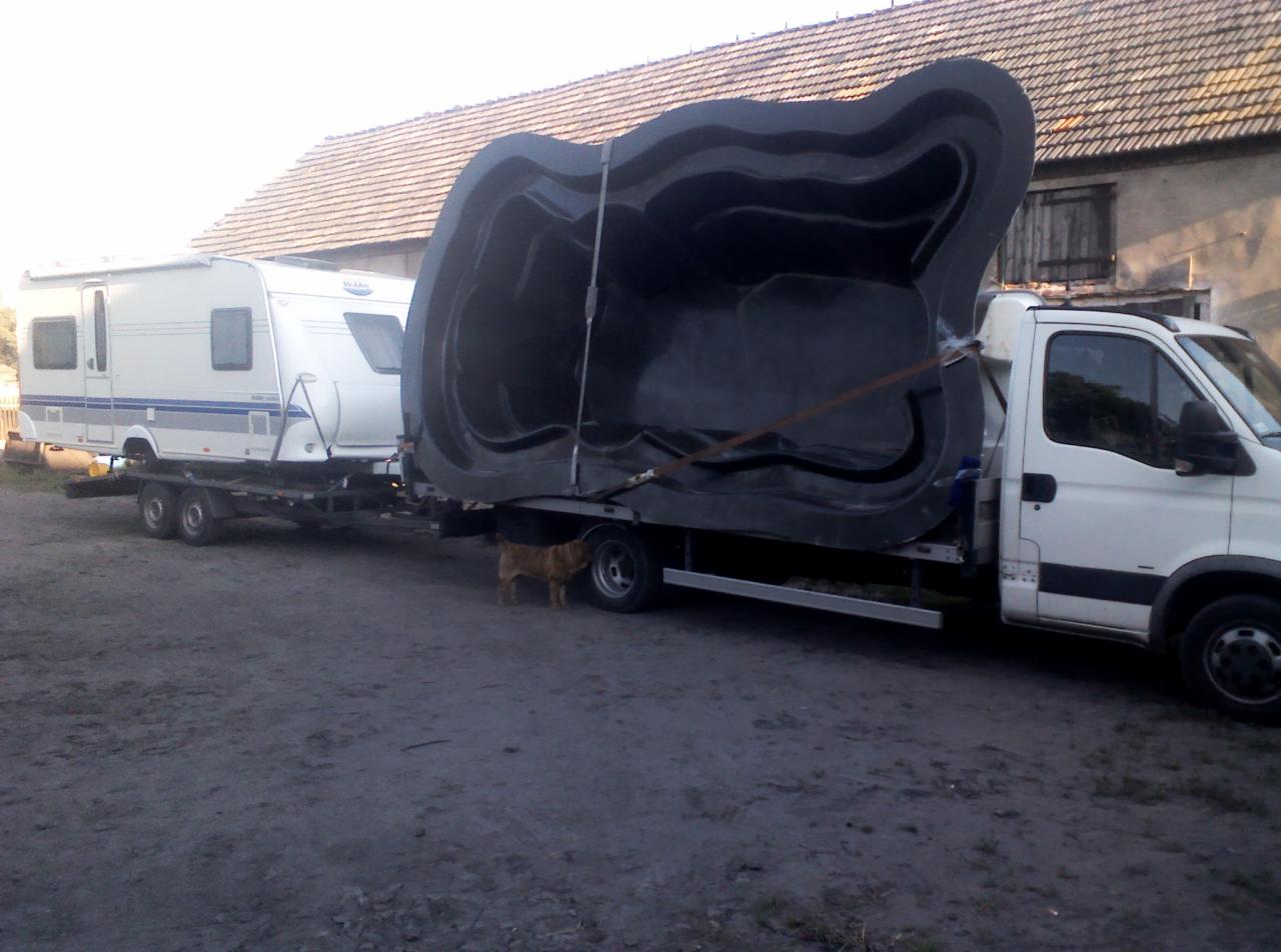 Transport przyczepy kempingowej i wanny basenowej na lawecie. Pies stoi pod lawetą. Budynek w tle.