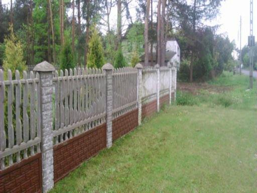 Szary, drewniany płot z betonowymi słupkami i brązową podmurówką z imitacji cegły, widoczny z perspektywy trawnika, z drzewami w tle.