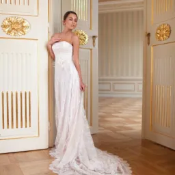 Elegancka kobieta w jasnej sukni stoi w otwartych, ozdobnych drzwiach z złoceniami, w tle widoczny klasyczny wystrój wnętrza z parkietem i pasami na ścianie.