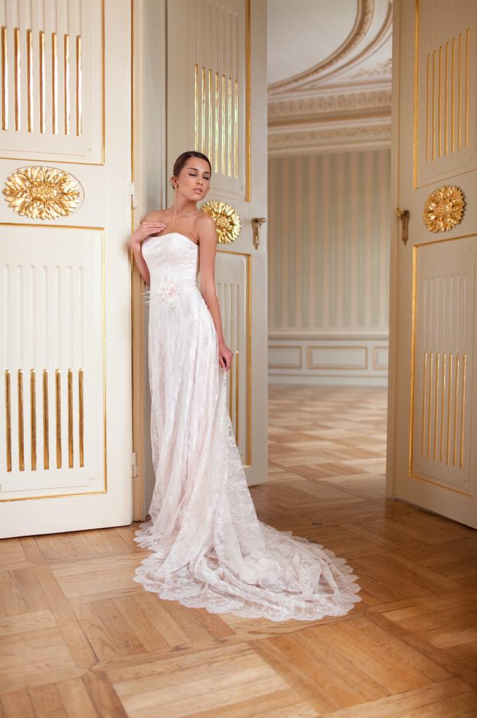 Elegancka kobieta w jasnej sukni stoi w otwartych, ozdobnych drzwiach z złoceniami, w tle widoczny klasyczny wystrój wnętrza z parkietem i pasami na ścianie.