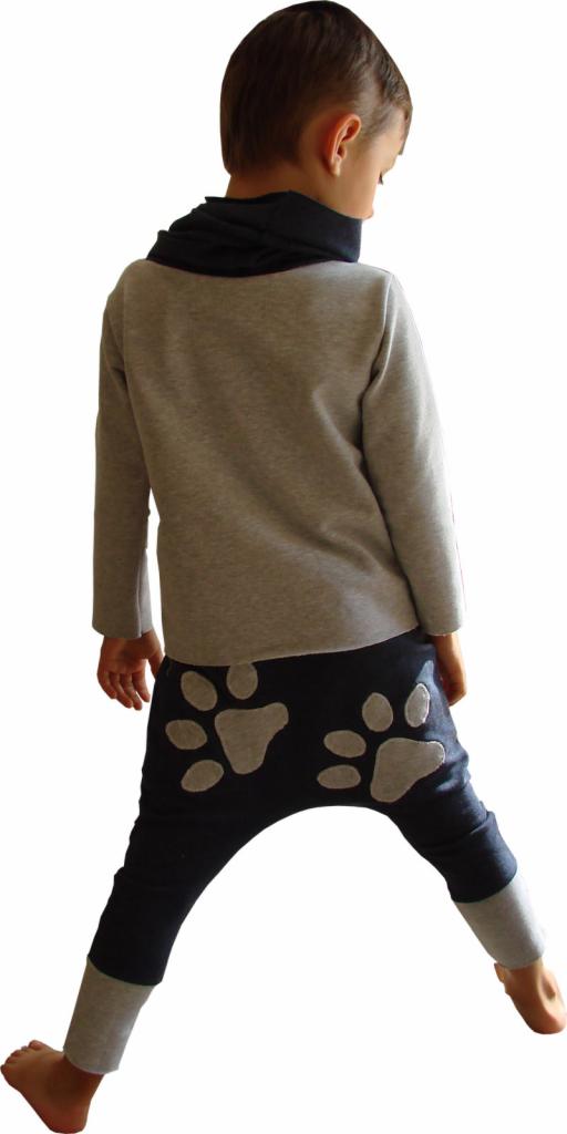 Chłopiec tyłem w szarej bluzce i ciemnych spodniach z aplikacjami łap. Widoczne szare mankiety spodni i czarny komin na szyi. Stonowana kolorystyka.