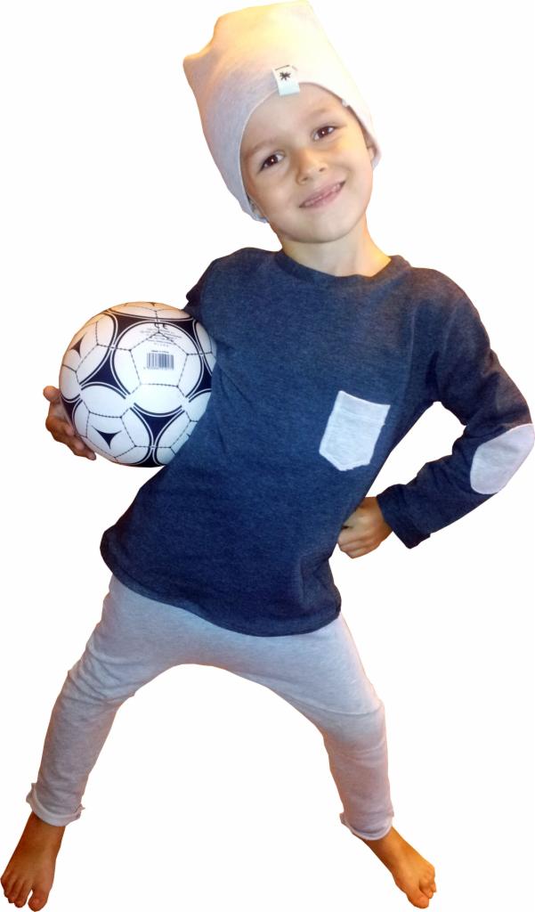 Uśmiechnięty chłopiec w szarej czapce i granatowej bluzce z łatami trzyma piłkę. Dziecięca stylizacja z kieszonką na bluzce i jasnymi spodniami.