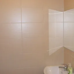 Beżowe płytki ścienne w łazience, narożne lustro z dwoma skrzydłami, umywalka z baterią i dozownikiem mydła w płynie.