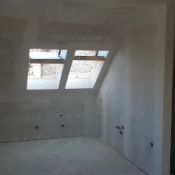 Pokój na poddaszu w trakcie remontu, ściany pokryte szpachlą, widoczne dwa okna dachowe, instalacja elektryczna wyprowadzona ze ścian, surowy stan podłogi.