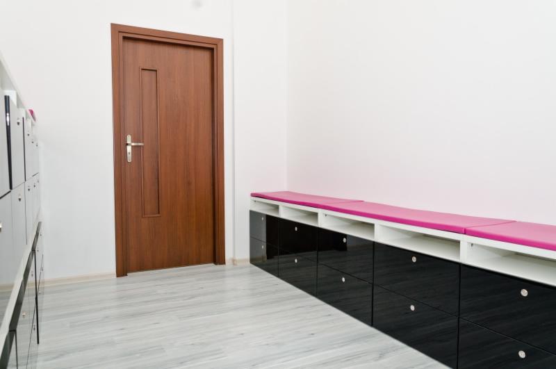 Nowoczesne wnętrze: drewniane drzwi z metalową klamką, szare panele podłogowe i szafka z różowym siedziskiem na tle białej ściany. Widoczne szafki po lewej stronie.