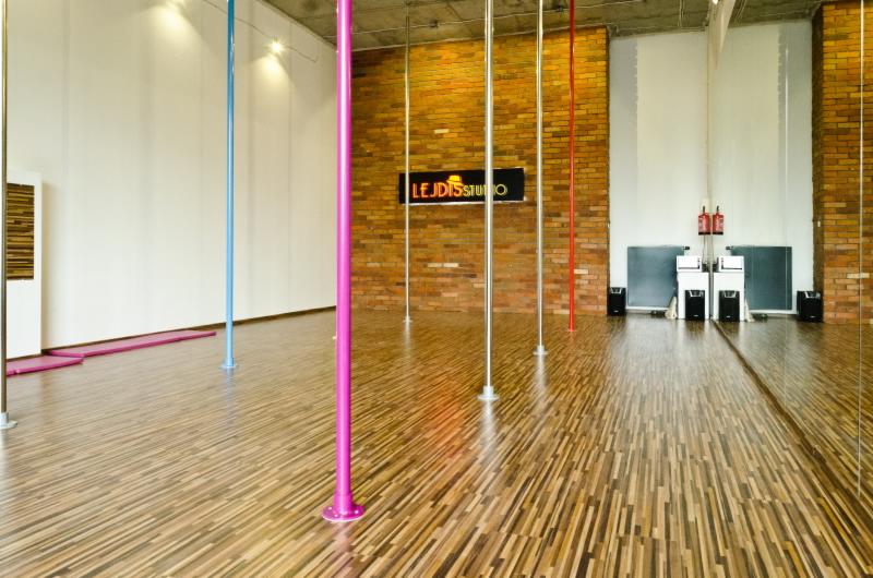 Sala do ćwiczeń z parkietem i kolorowymi rurkami do pole dance. Na ścianie logo studia na tle cegieł. Lustro odbija przestrzeń. Widok na całe pomieszczenie.
