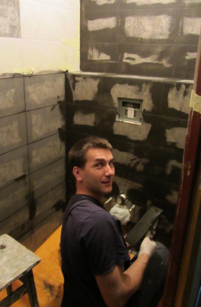 Mężczyzna w trakcie fugowania płytek w łazience. Widoczne narzędzia i częściowo wykończone ściany. Praca w toku, z uśmiechem na twarzy.