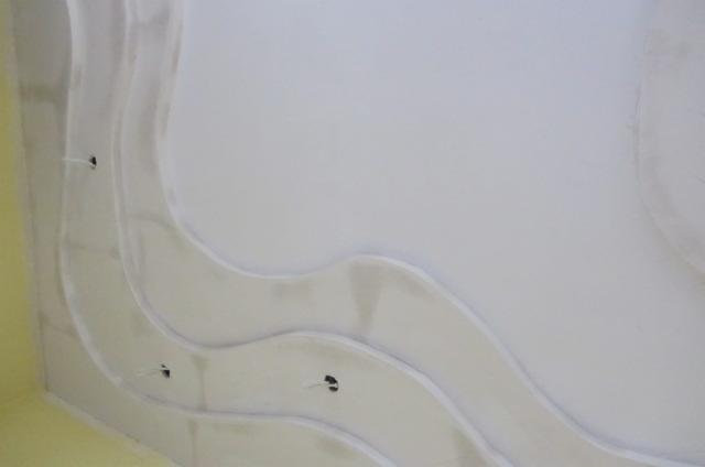 Biały sufit z dekoracyjnymi, gipsowymi zdobieniami w kształcie fal, widoczne otwory na oświetlenie. Minimalistyczny design.