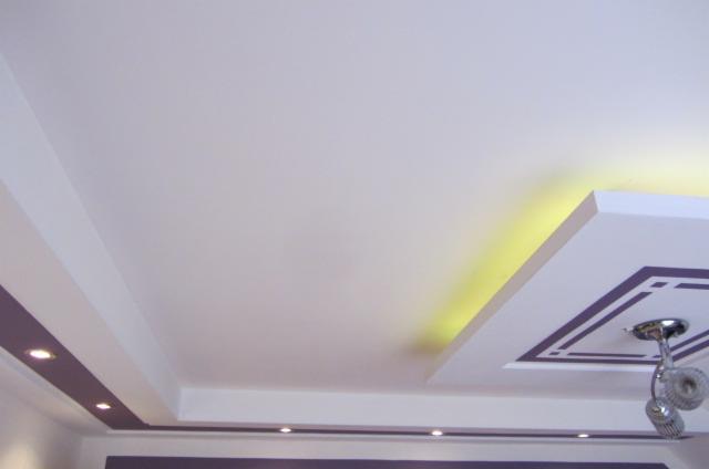 Biały sufit z podwieszanymi elementami i oświetleniem punktowym oraz dekoracyjnym, fioletowe akcenty.