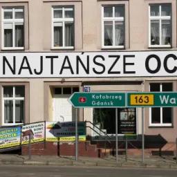 Budynek z szyldem 'Najtańsze OC' i reklamami ubezpieczeń: na życie, mieszkania i domów. Tablice kierunkowe do Kołobrzegu i Wałcza. Reklama firmy ubezpieczeniowej.