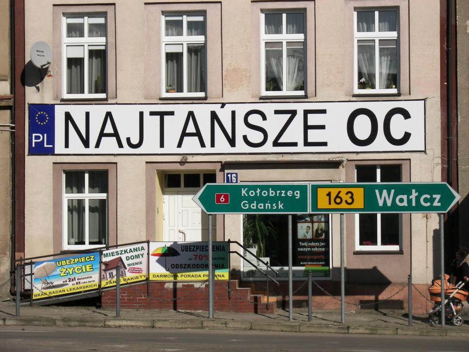 Budynek z szyldem 'Najtańsze OC' i reklamami ubezpieczeń: na życie, mieszkania i domów. Tablice kierunkowe do Kołobrzegu i Wałcza. Reklama firmy ubezpieczeniowej.
