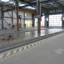 Posadzka przemysłowa w hali produkcyjnej z żółto-czarnymi pasami ostrzegawczymi. Widoczne narzędzia i osoba w oddali. Duże okna w tle.