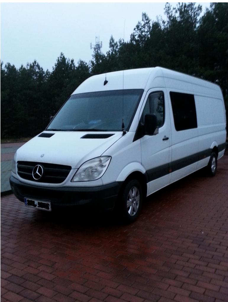 Biały, dostawczy Mercedes Sprinter na czerwonej kostce brukowej, tle lasu i masztu telekomunikacyjnego. Widok z przodu, lekko z boku. Szyba boczna częściowo zasłonięta.