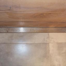 INWK sp. z o. o. - Nowoczesny odpływ liniowy z perforowaną maskownicą wkomponowany w szarą, kwadratową glazurę podłogową i drewnopodobną okładzinę ścienną w kabinie prysznicowej.