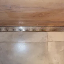 Nowoczesny odpływ liniowy z perforowaną maskownicą wkomponowany w szarą, kwadratową glazurę podłogową i drewnopodobną okładzinę ścienną w kabinie prysznicowej.