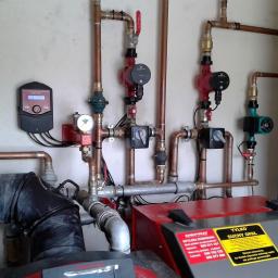 INWK sp. z o. o. - Widok z bliska instalacji grzewczej z kotłem Defro, rurami miedzianymi, pompami obiegowymi Grundfos i sterownikiem, z naklejkami informacyjnymi na kotle.