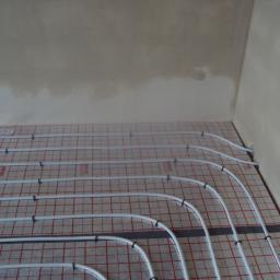 INWK sp. z o. o. - Białe rury ogrzewania podłogowego ułożone na folii z czerwoną kratką, przymocowane czarnymi klipsami do podłoża w rogu pomieszczenia przed wylaniem posadzki.