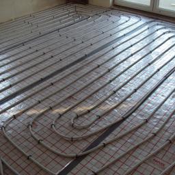 INWK sp. z o. o. - Równolegle ułożone, białe rury ogrzewania podłogowego na folii z czerwoną kratką, zabezpieczone czarnymi uchwytami, widoczne okno w tle.