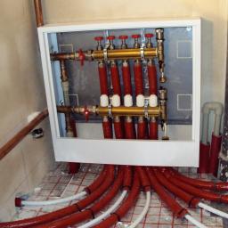 INWK sp. z o. o. - Rozdzielacz ogrzewania podłogowego w białej obudowie, z czerwonymi rurami wychodzącymi w dół, ułożonymi na folii z siatką, widoczna część instalacji miedzianej na ścianie.