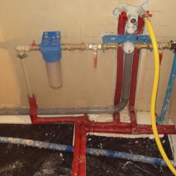 INWK sp. z o. o. - Węzeł instalacyjny z filtrem wody, rurami czerwonymi, szarymi i niebieskimi, oraz żółtym przewodem gazowym, zamontowany przy ścianie i podłodze w trakcie prac wykończeniowych.