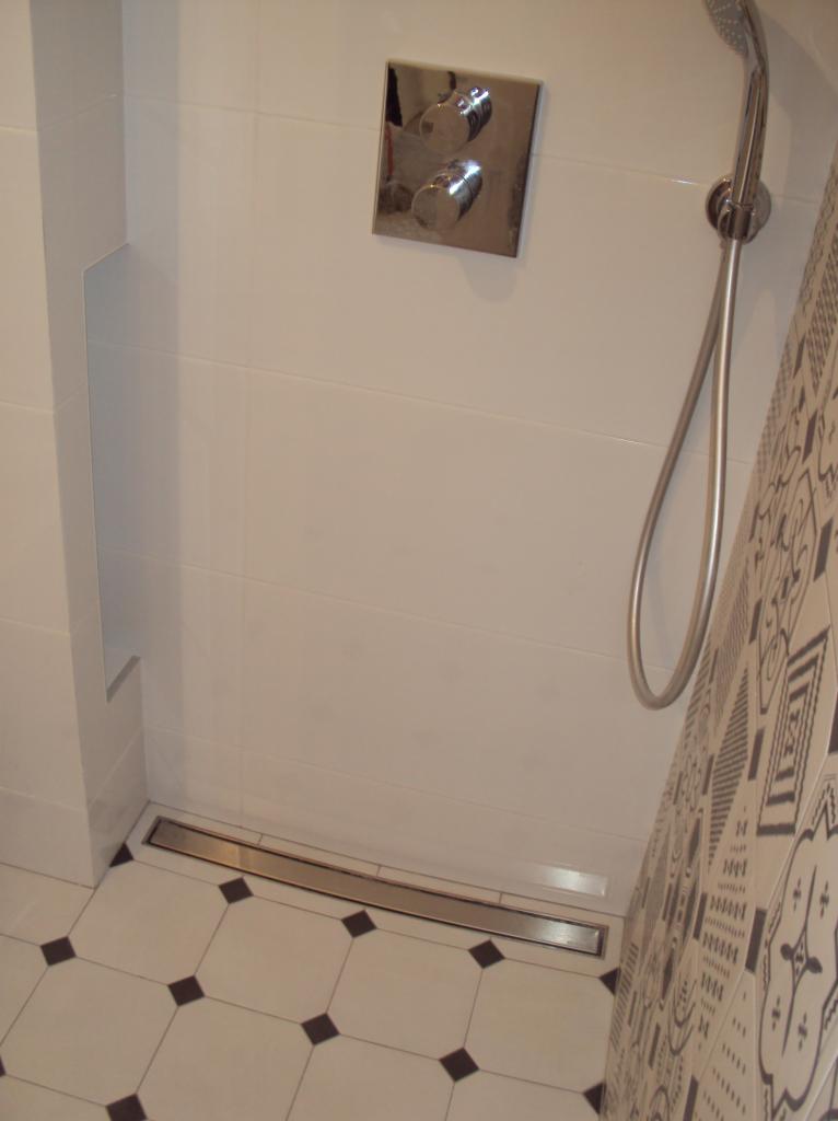 Nowoczesna kabina prysznicowa z odpływem liniowym i chromowaną armaturą, wykończona białymi płytkami z czarnymi akcentami na podłodze.