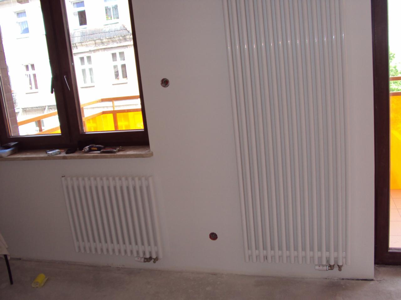 Białe, pionowe grzejniki żeberkowe na ścianie w pokoju. Jeden pod oknem, drugi obok drzwi balkonowych. Widoczne otwory w ścianie na instalacje.