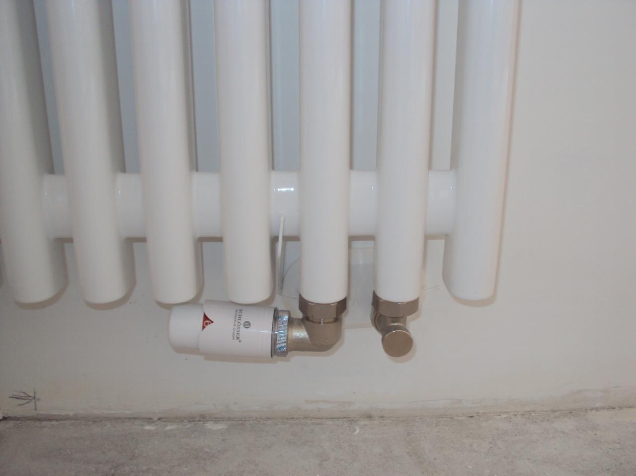 Biały, żeliwny grzejnik żeberkowy z zamontowanym termostatem Schlosser, widok z dołu, na tle jasnej ściany. Detal instalacji grzewczej.