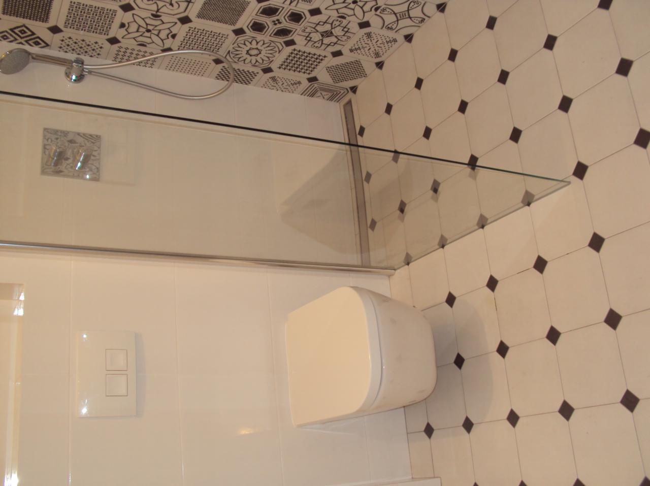 Nowoczesna łazienka z kabiną prysznicową i podwieszaną toaletą, wykończona białymi płytkami z ciemnymi akcentami i dekoracyjnym wzorem na suficie.