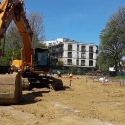 Żółta koparka na placu budowy, przygotowującym teren pod fundamenty, widoczne pionowe rury szalunkowe, w tle budynki mieszkalne i pracownicy w kamizelkach odblaskowych.