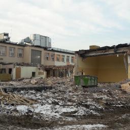 P.H.U. RAVI TRANS - Częściowo zburzony budynek szkoły z widocznym gruzem, stertą drewna, kontenerem i koparką w tle, teren pokryty błotem i resztkami śniegu.