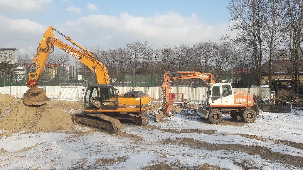 Dwie koparki, żółta CASE CX240 i pomarańczowa ATLAS 1504, pracują na zasypanym śniegiem placu budowy; w tle budynki i drzewa bez liści.