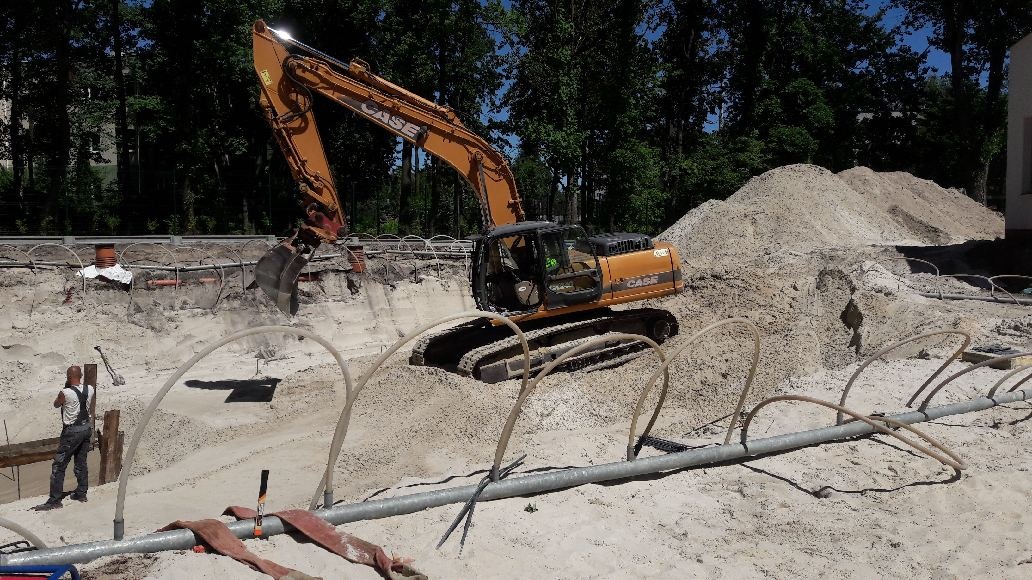 Pomarańczowa koparka Case wykonuje wykop w piaszczystym gruncie, widoczne rury drenażowe i pracownik w tle.