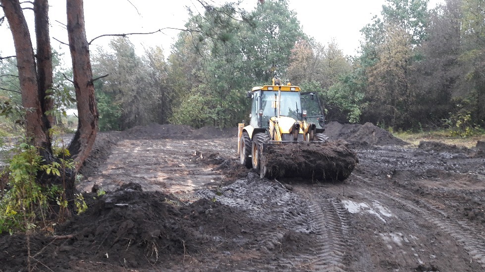 Żółta koparko-ładowarka utwardza błotnistą drogę gruntową na tle drzew, widoczne ślady opon i świeżo rozplantowana ziemia.