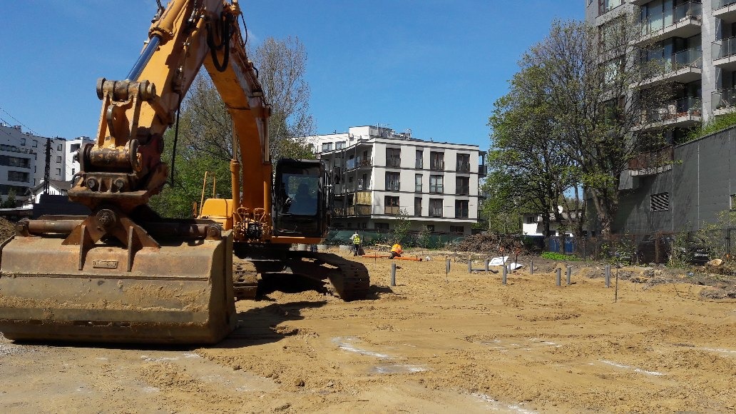 Żółta koparka na placu budowy, przygotowującym teren pod fundamenty, widoczne pionowe rury szalunkowe, w tle budynki mieszkalne i pracownicy w kamizelkach odblaskowych.