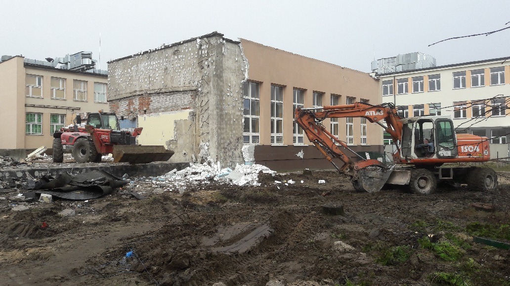 Pomarańczowa koparka ATLAS 1504 i czerwony ładowacz teleskopowy pracują na placu budowy z gruzem, w tle budynki szkolne.