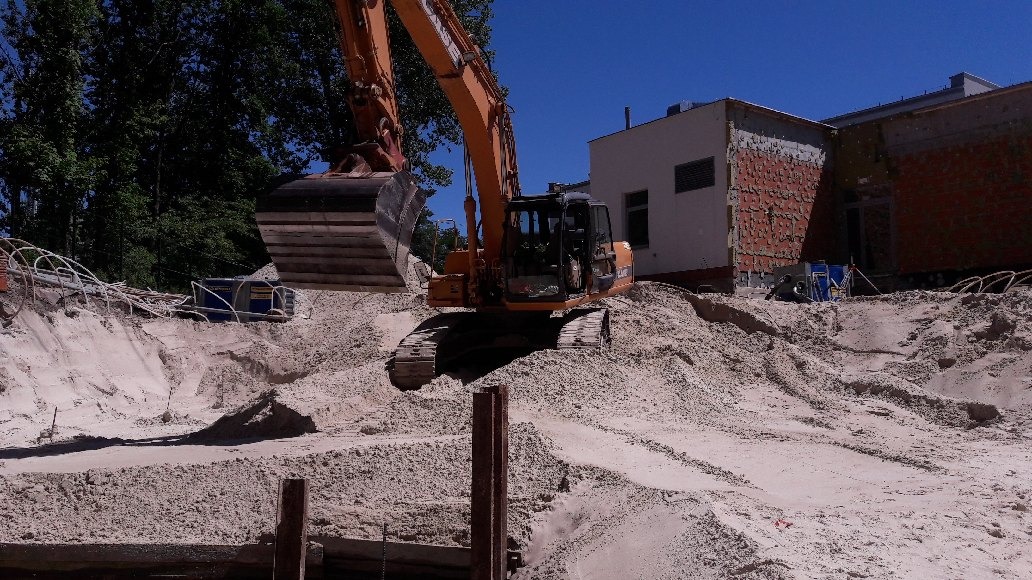 Pomarańczowy koparka gąsienicowa na placu budowy, w tle budynek w trakcie wznoszenia, wykop wypełniony piaskiem.