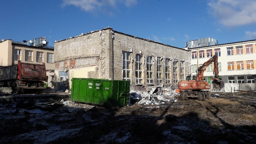 Pomarańczowa koparka Atlas 1504 i czerwona ciężarówka wywożą gruz ze zburzonego budynku z zielonym kontenerem EKO RAFF w tle pod błękitnym niebem.