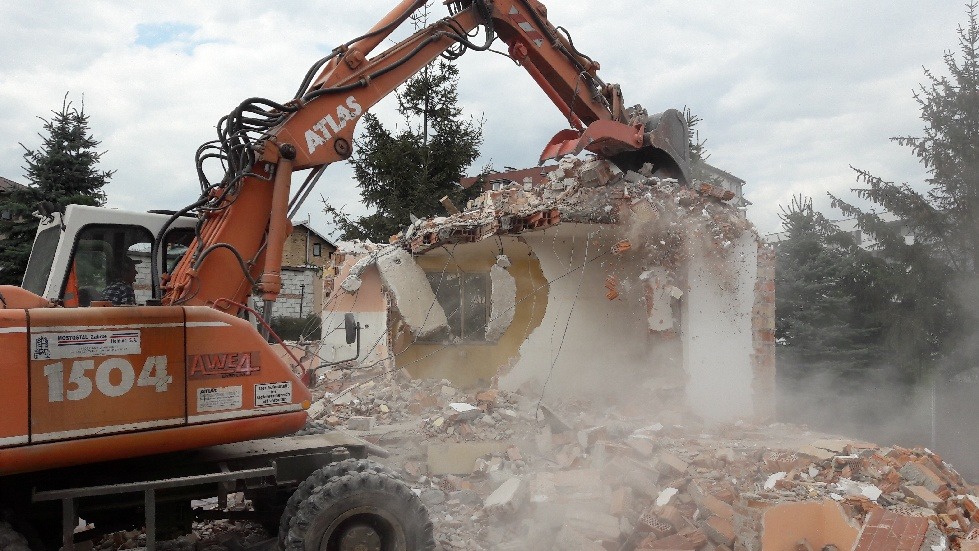 Pomarańczowa koparka Atlas 1504 kruszy fragment budynku z cegły i betonu. Na pierwszym planie gruz i kurz, w tle drzewa i budynki.