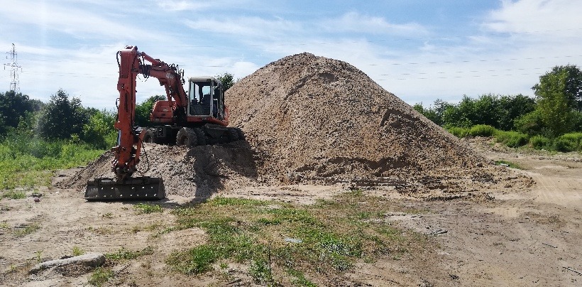 Pomarańczowa koparka kołowa stoi obok dużej pryzmy ziemi na placu budowy, w tle widoczne drzewa i słup wysokiego napięcia.