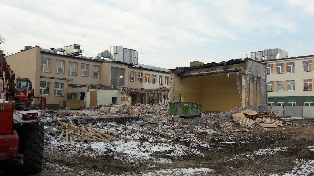 Częściowo zburzony budynek szkoły z widocznym gruzem, stertą drewna, kontenerem i koparką w tle, teren pokryty błotem i resztkami śniegu.