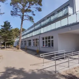 Zewnętrzny widok rozbudowanego, trzypiętrowego ośrodka rehabilitacyjno-leczniczego z balkonami z mlecznego szkła, z rampą dla niepełnosprawnych prowadzącą do wejścia, otoczonego drzewami iglastymi...