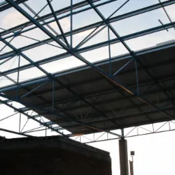 Wnętrze hali produkcyjnej w trakcie budowy, widok na konstrukcję stalową dachu z niebieskimi elementami, betonowe słupy podtrzymujące konstrukcję oraz fragment muru z cegły w dolnej części kadru.