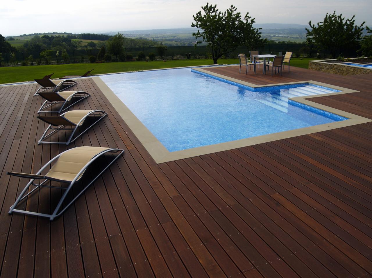 Nowoczesny basen z ciemnobrązowym tarasem z desek, leżaki z beżowymi siedziskami ustawione wzdłuż krawędzi, w tle zielony krajobraz z drzewami i wzgórzami.