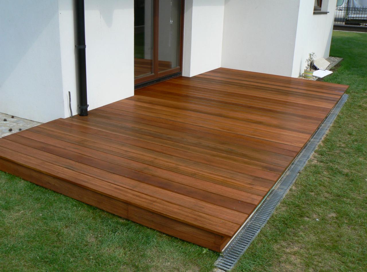 Nowoczesny taras z egzotycznego drewna, z subtelnym spadkiem i odpływem liniowym, przylegający do białej ściany budynku z brązowymi oknami, otoczony zielonym trawnikiem.