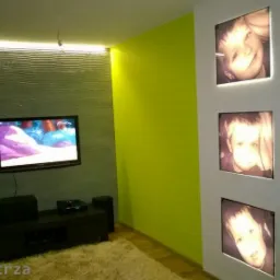Żywy, limonkowy akcent na ścianie kontrastuje z szarą, teksturowaną ścianą TV. Trzy podświetlane portrety dziecięce zdobią białą kolumnę w nowoczesnym wnętrzu.