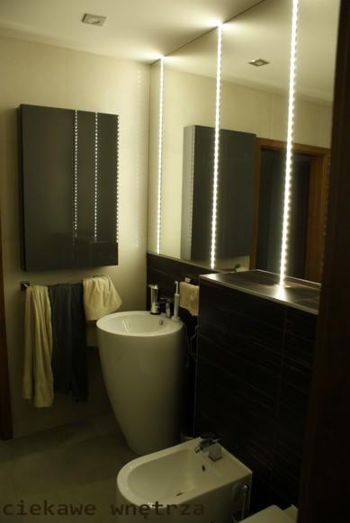 Nowoczesna łazienka z podświetlanym lustrem, umywalką na postumencie i bidetem. Ciemne płytki i jasne ściany. Widoczne ręczniki i szafka z lustrem.