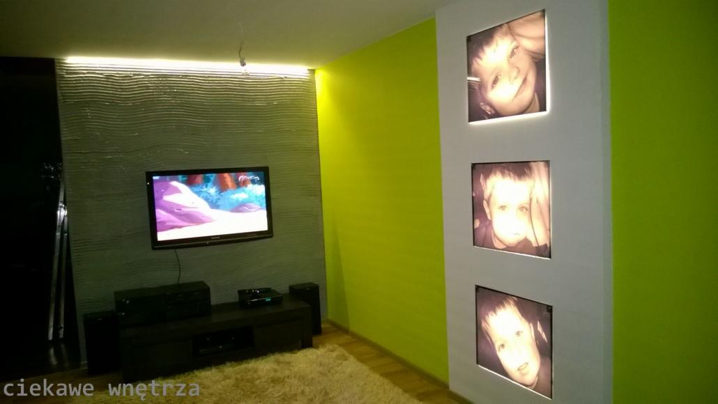 Żywy, limonkowy akcent na ścianie kontrastuje z szarą, teksturowaną ścianą TV. Trzy podświetlane portrety dziecięce zdobią białą kolumnę w nowoczesnym wnętrzu.
