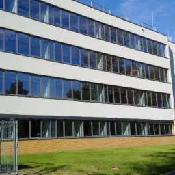 Elewacja budynku biurowego z rzędami okien odbijających niebo, widoczny trawnik i fragment ogrodzenia. Minimalistyczna architektura, jasne tynki zewnętrzne.